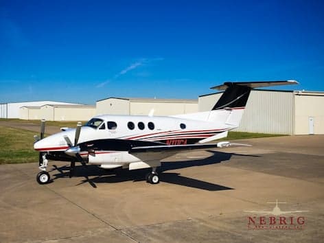 A white Beechcraft King Air twin-turboprop with black and red stripes and a black tail, parked on an airport tarmac with hangars in the background under a clear blue sky.