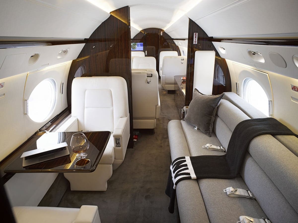 A long view of the Gulfstream G650 cabin interior, featuring white leather seats, a grey sofa, and dark wood accents.