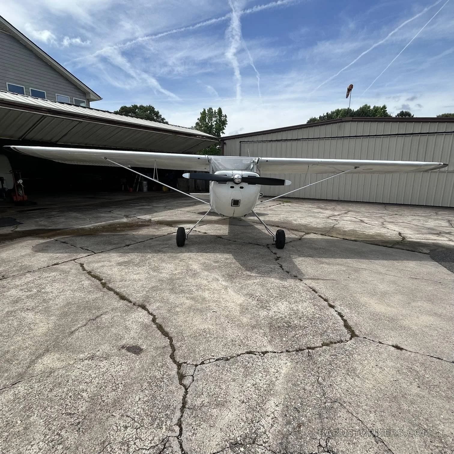 A white Cessna 170B with dark blue and gold stripes is viewed head-on, parked on a concrete pad in front of a hangar.