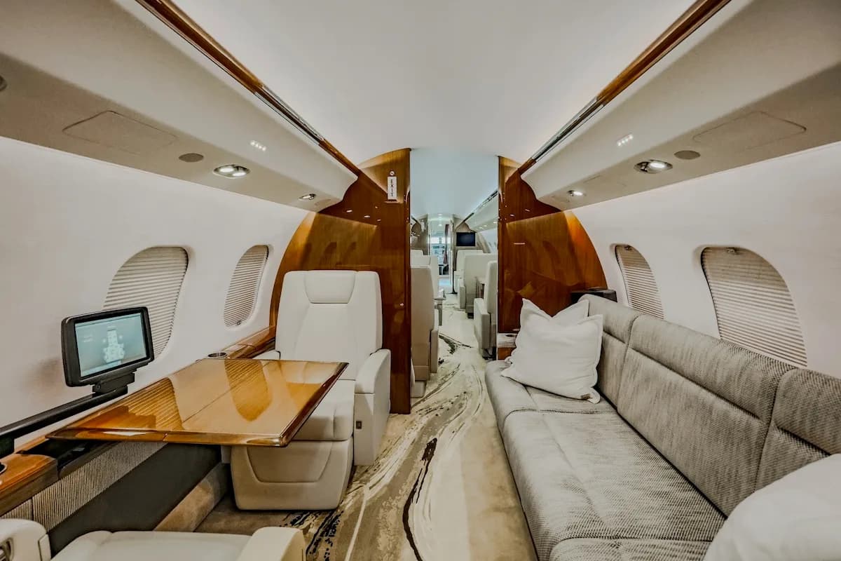 A private jet cabin interior featuring a long couch on one side and individual seats with a table and monitor on the other.