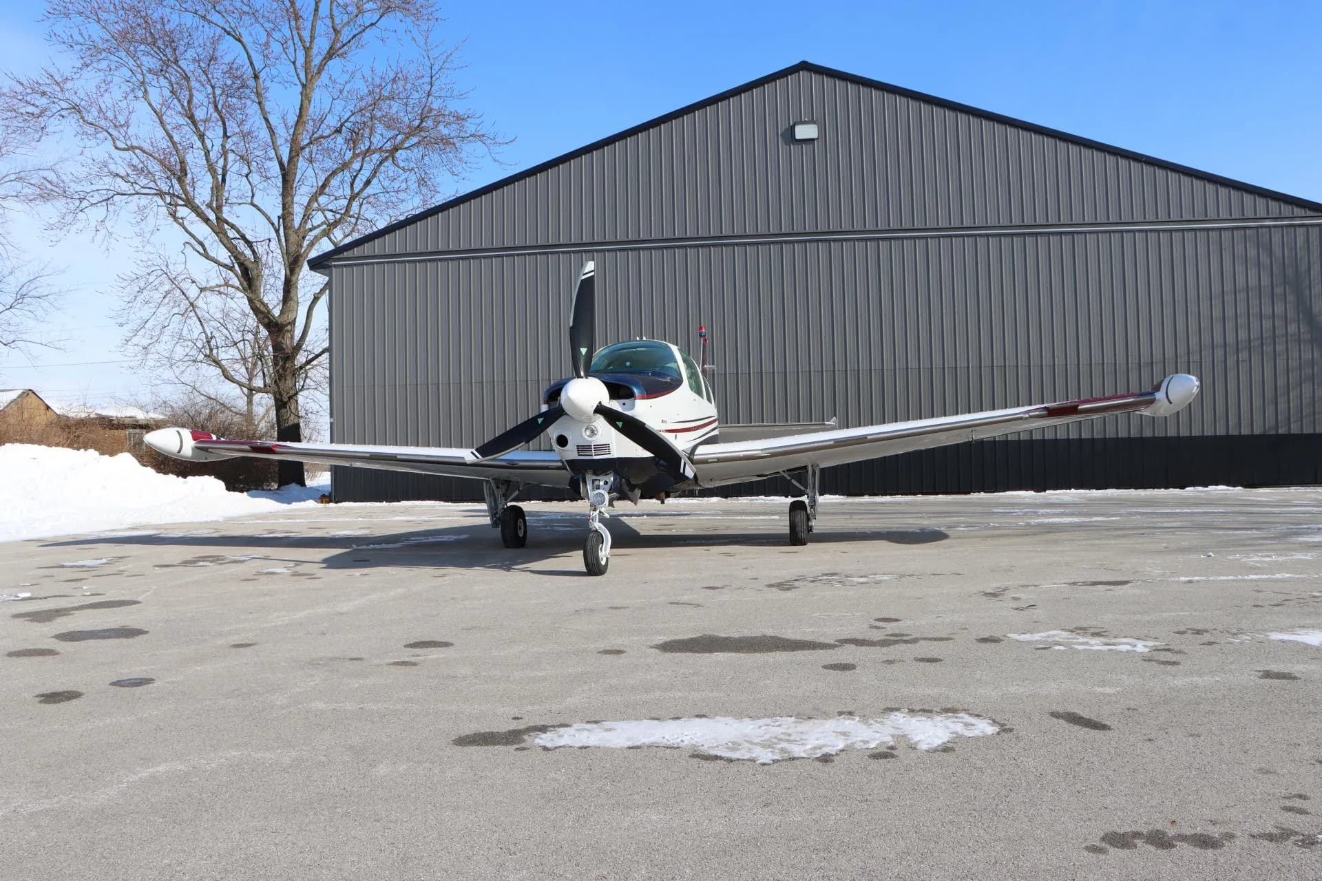 Front view of a white, blue, and red single-engine aircraft parked on a partially snowy tarmac in front of a hangar and bare trees.