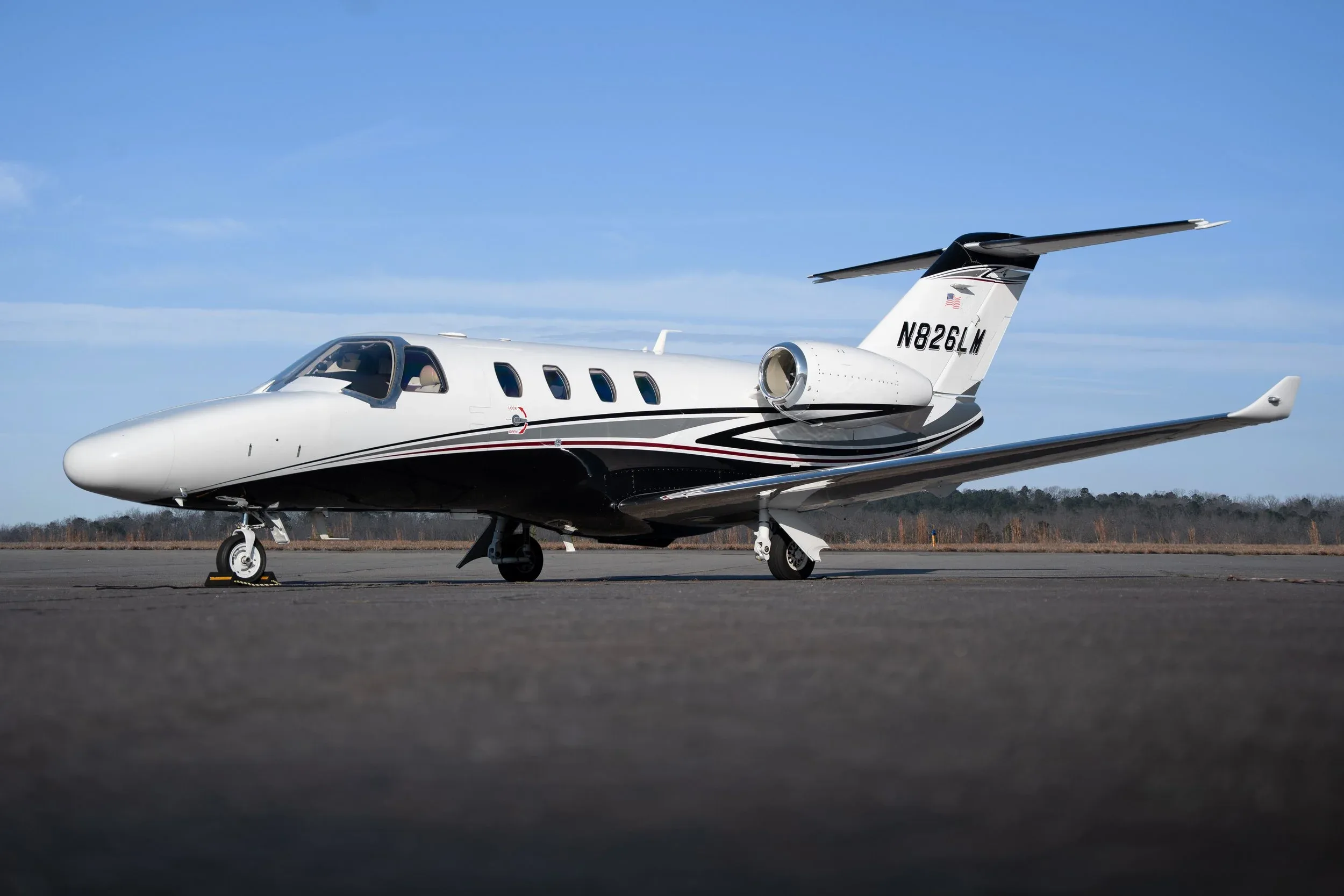 A white business jet with black and red stripes, registration N826LM, parked on an asphalt runway under a clear blue sky.