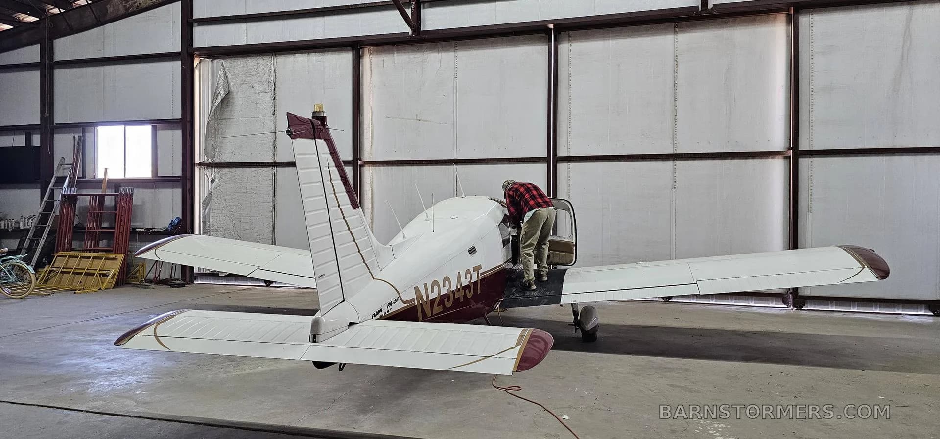 The rear view of a white and maroon Piper Cherokee 140 (N9243T) inside a hangar, with a person leaning into the cockpit.