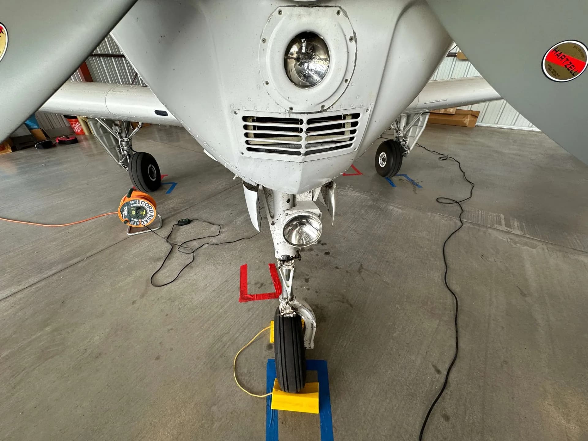 A view from directly under the nose of a Beechcraft Bonanza, showing the nose landing gear, propeller blades, and a ground power unit.