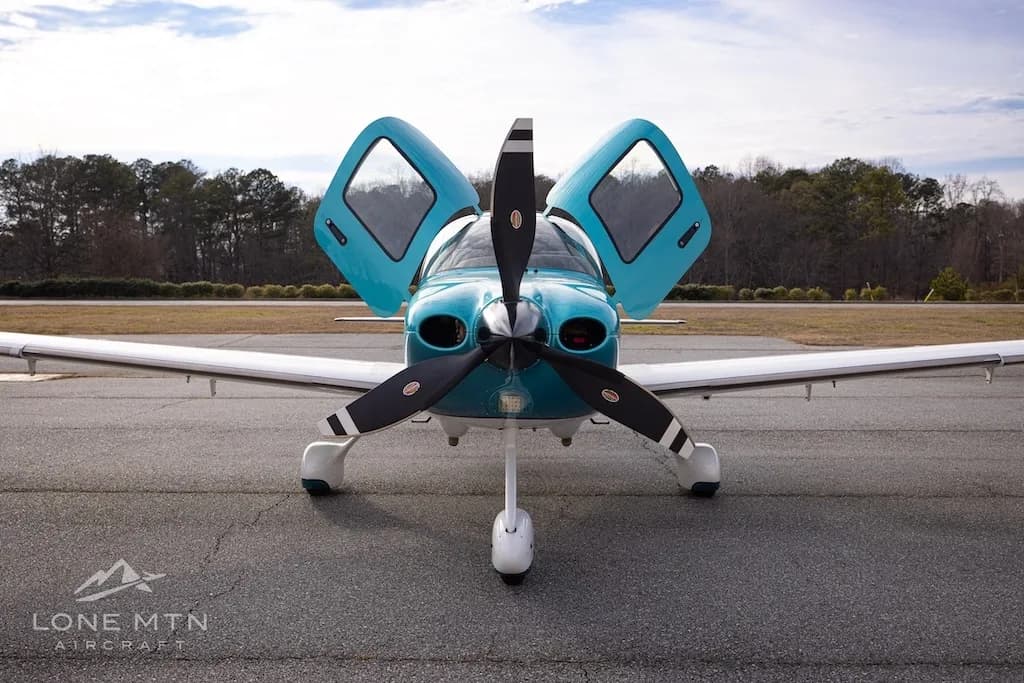 A head-on view of the teal and white Cirrus SR22 aircraft, N908DU, with its doors open, parked on an airfield.