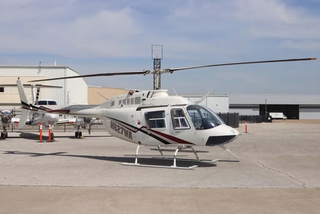 A white Bell 206 helicopter with N827WA registration on the side, parked on an airfield with hangars in the background.