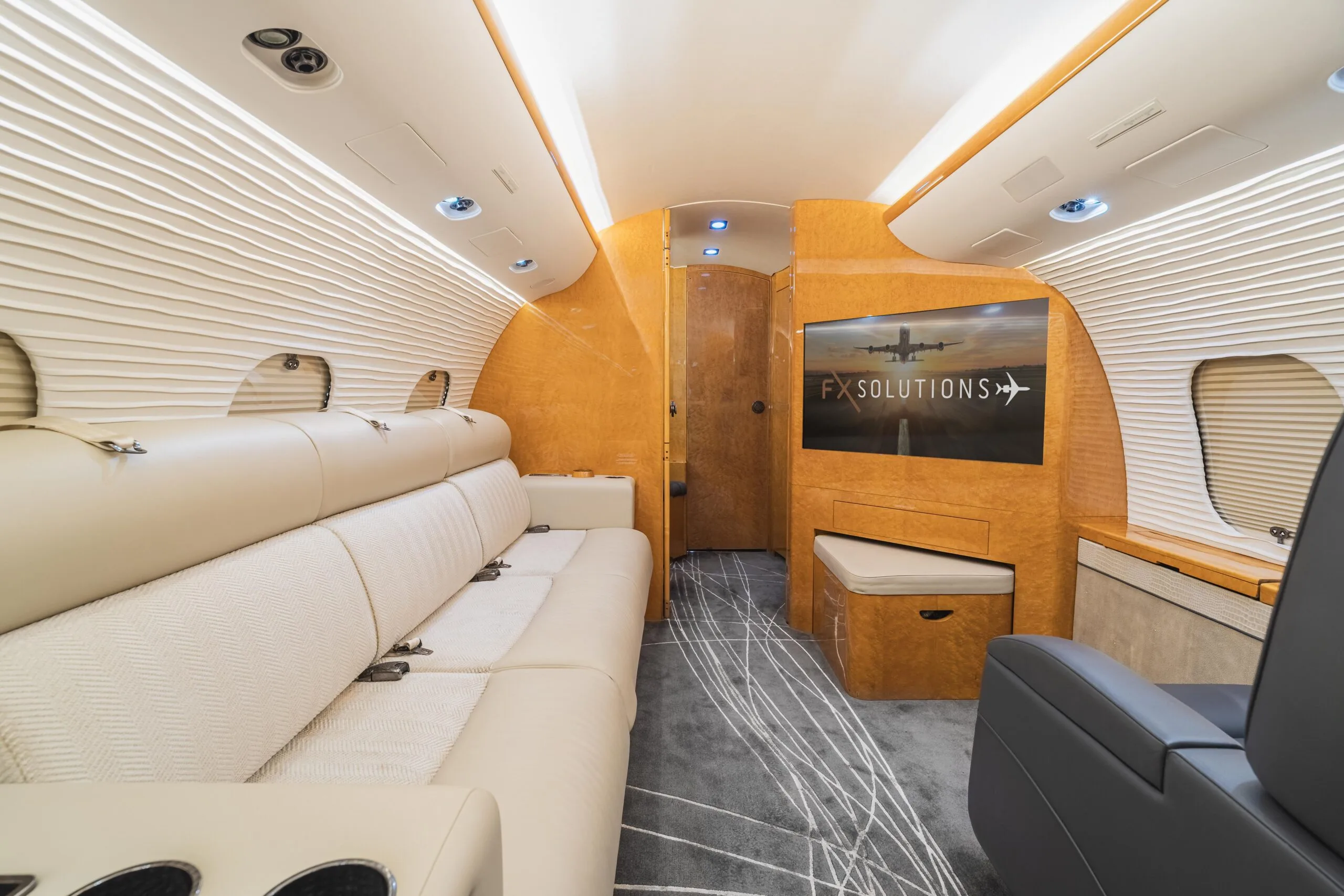 A view of the business jet cabin interior, showing a cream-colored sofa, a large entertainment screen displaying "FX SOLUTIONS+", and light wood paneling.