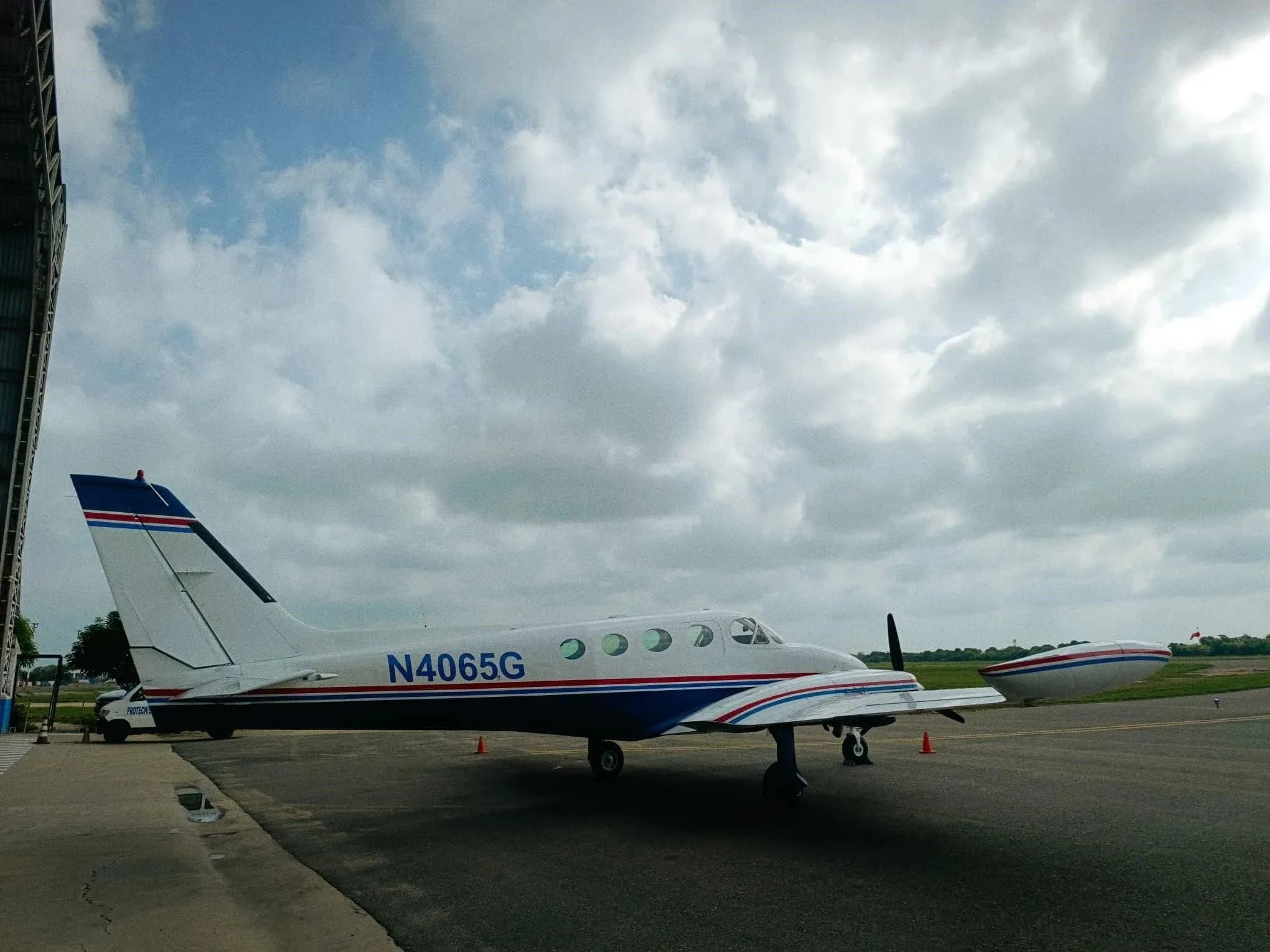 A side view of a white and blue Cessna 340 twin-engine aircraft with red stripes parked on an airfield next to a hangar.