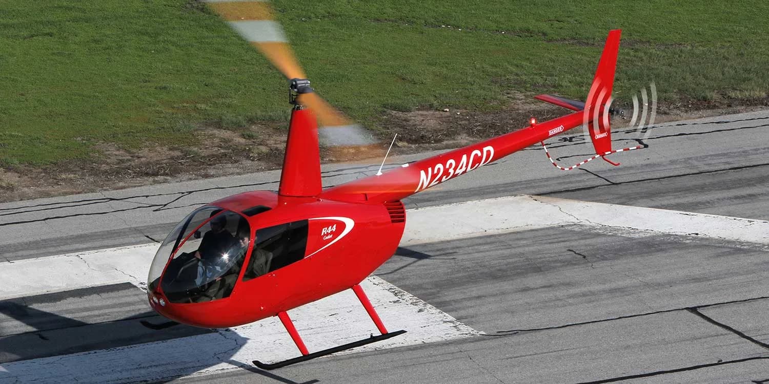 A red Robinson R44 helicopter taking off from a runway.