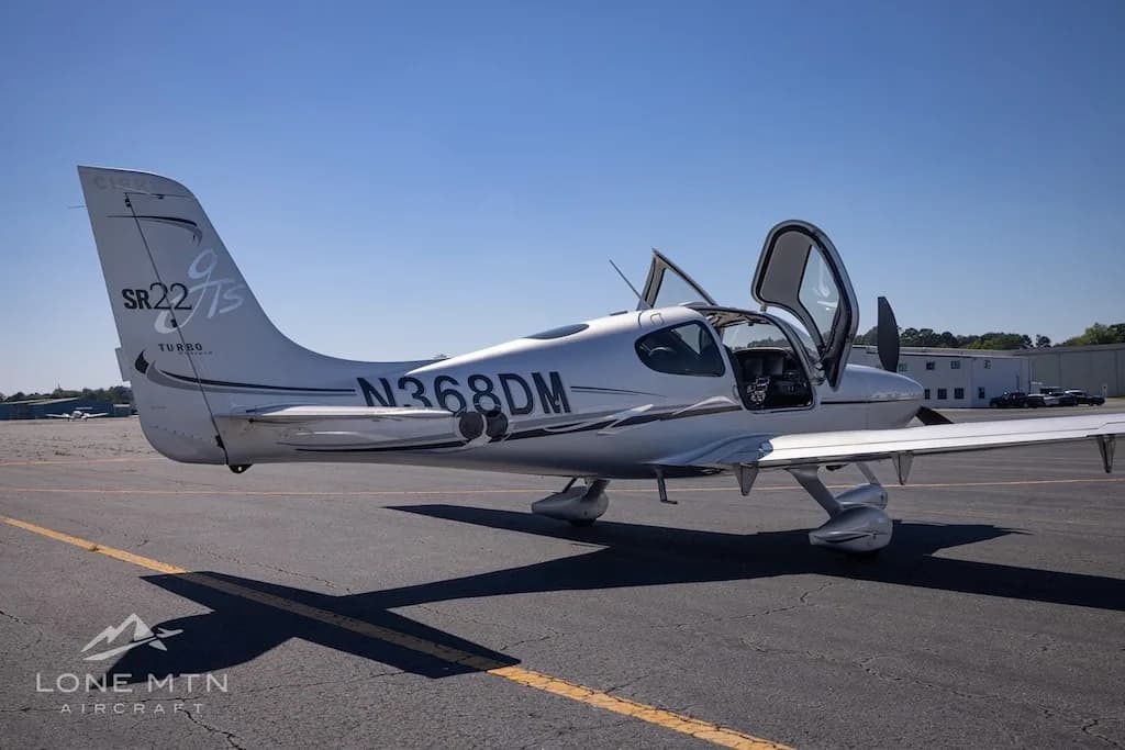 A silver Cirrus SR22 Turbo aircraft (N368DM) with its doors open, parked on an asphalt runway under a clear blue sky.