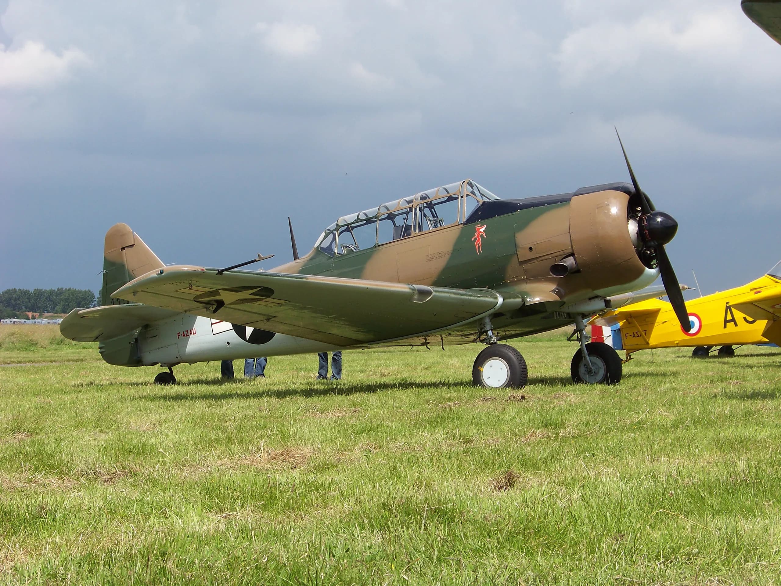 A North American T-6 Texan aircraft, F-AZAU, in camouflage paint scheme, parked on a grassy field under a cloudy sky.