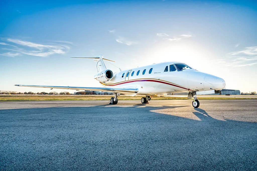 A white business jet with red and gold stripes (N24KT) is parked on a tarmac under a bright blue sky.