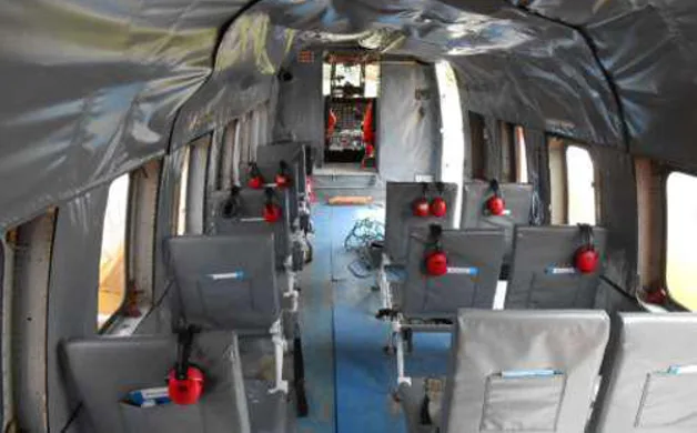 The interior cabin of a helicopter features rows of grey seats with red headphones, looking towards the cockpit.
