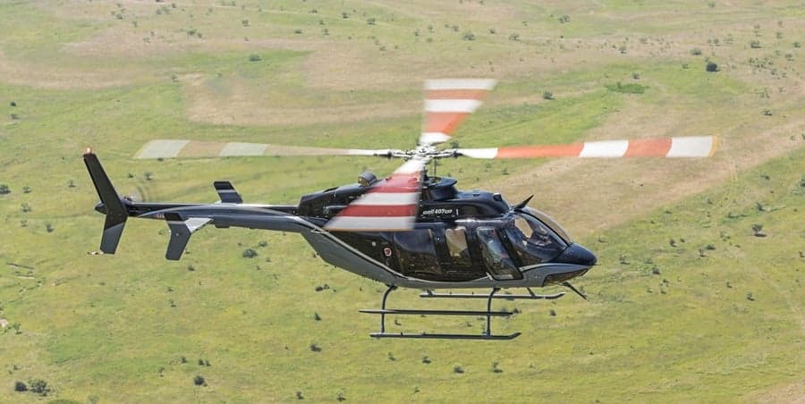 Bell 407 helicopter in flight over a grassy field.