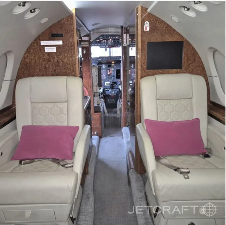 The interior of a private jet cabin looking forward, showing two light-colored leather seats with pink pillows and the entrance to the cockpit.