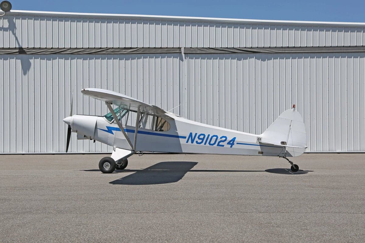 Side view of a white and blue Piper Cub in front of a hangar.