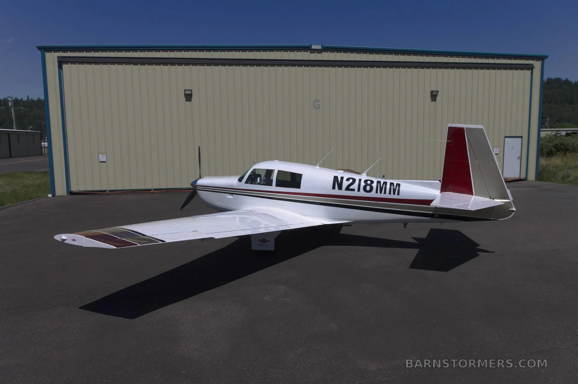 A white single-engine propeller aircraft with red and brown stripes, viewed from the rear-left, parked on an asphalt surface in front of a light-colored hangar.