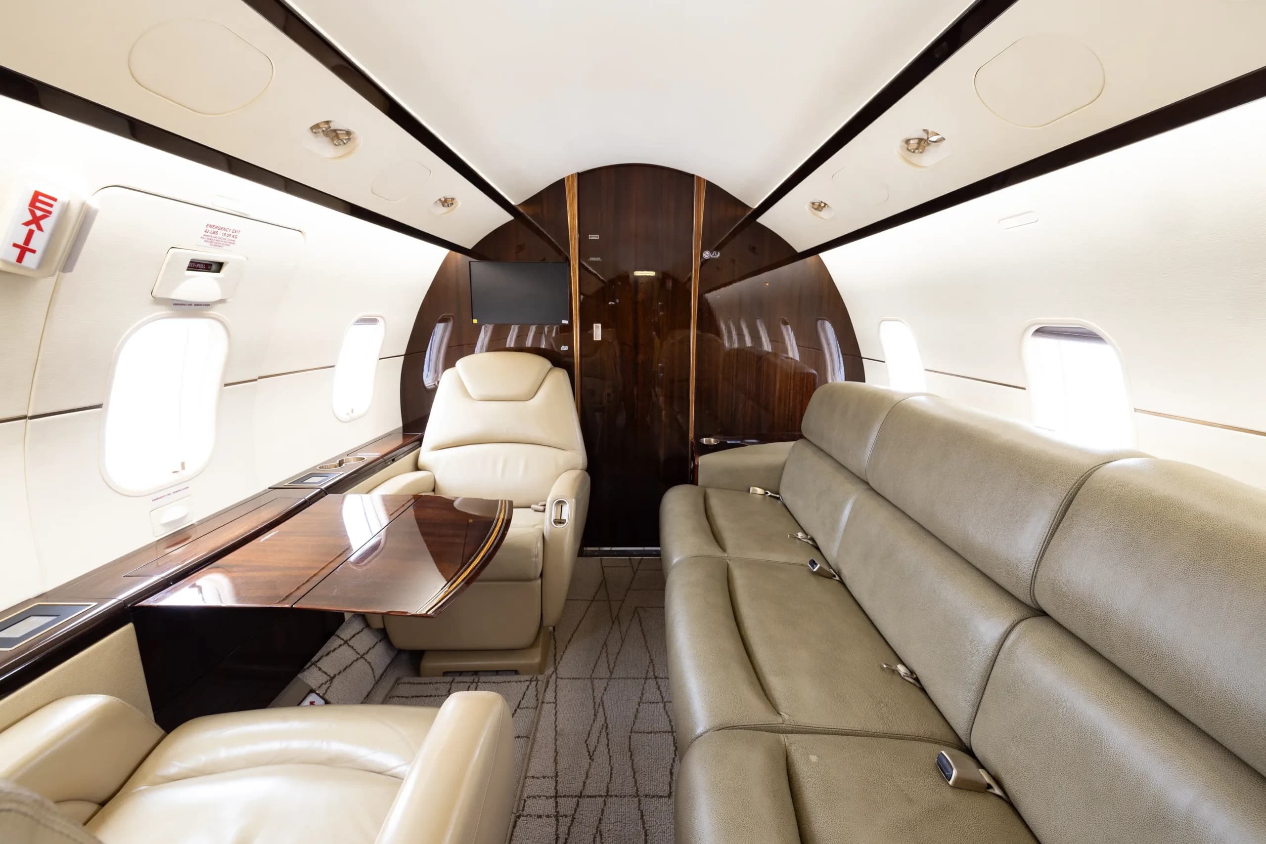 The rear section of a business jet cabin featuring a long beige leather sofa and a single seat with a wooden table.