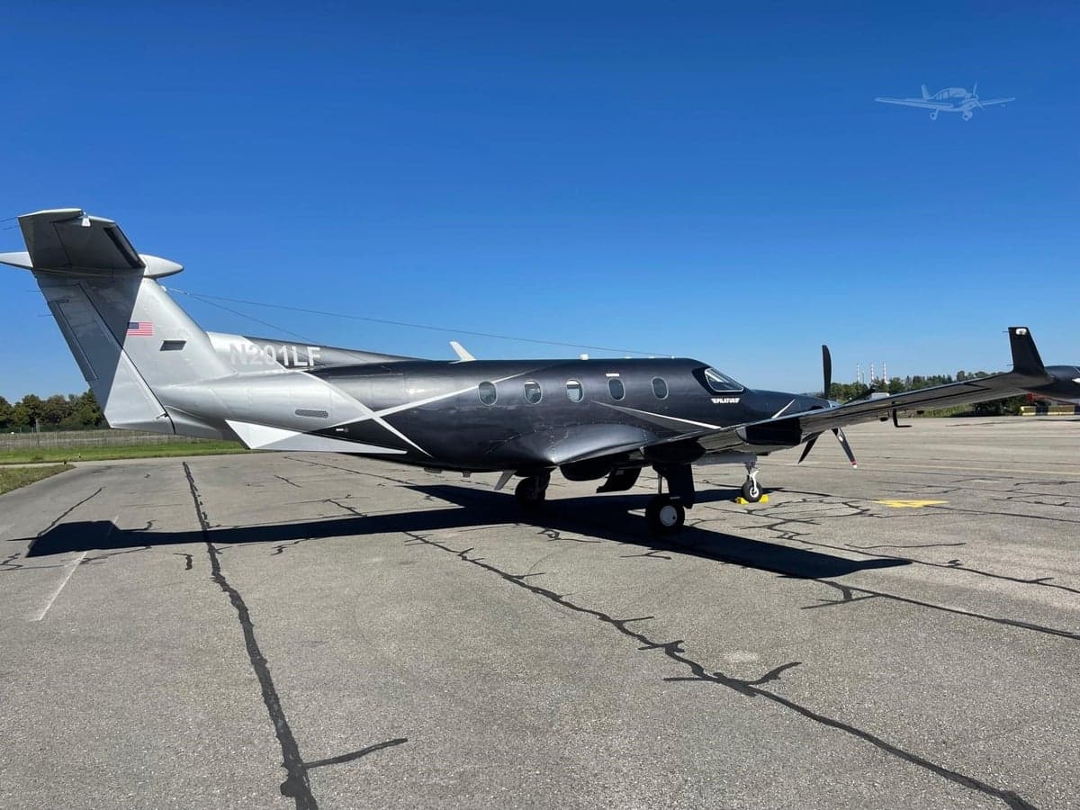 A dark grey and silver Pilatus PC-12 turboprop aircraft with white accents is parked on an asphalt tarmac, viewed from the rear-left.