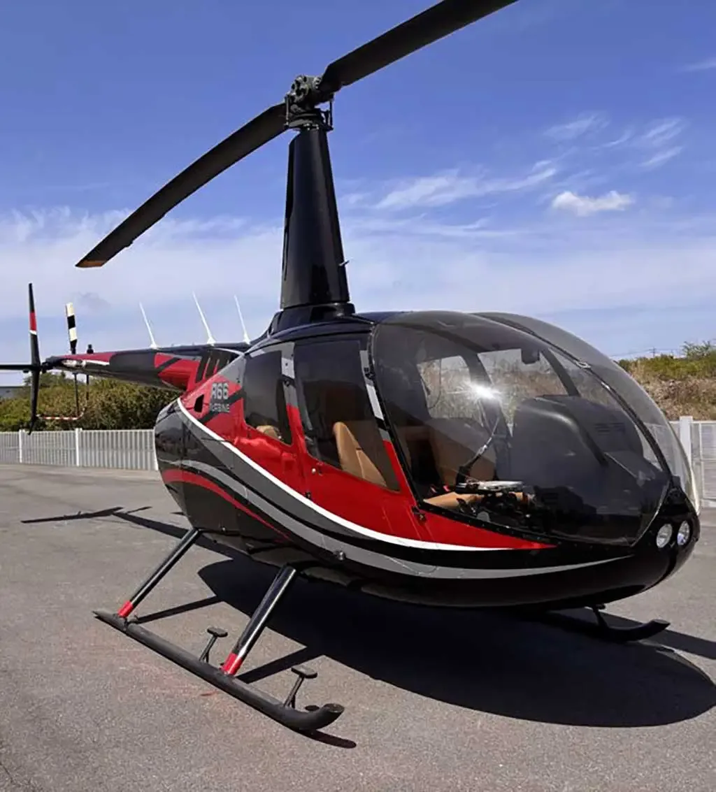 A black and red Robinson R66 Turbine helicopter with tan interior is parked on an asphalt surface under a blue sky.