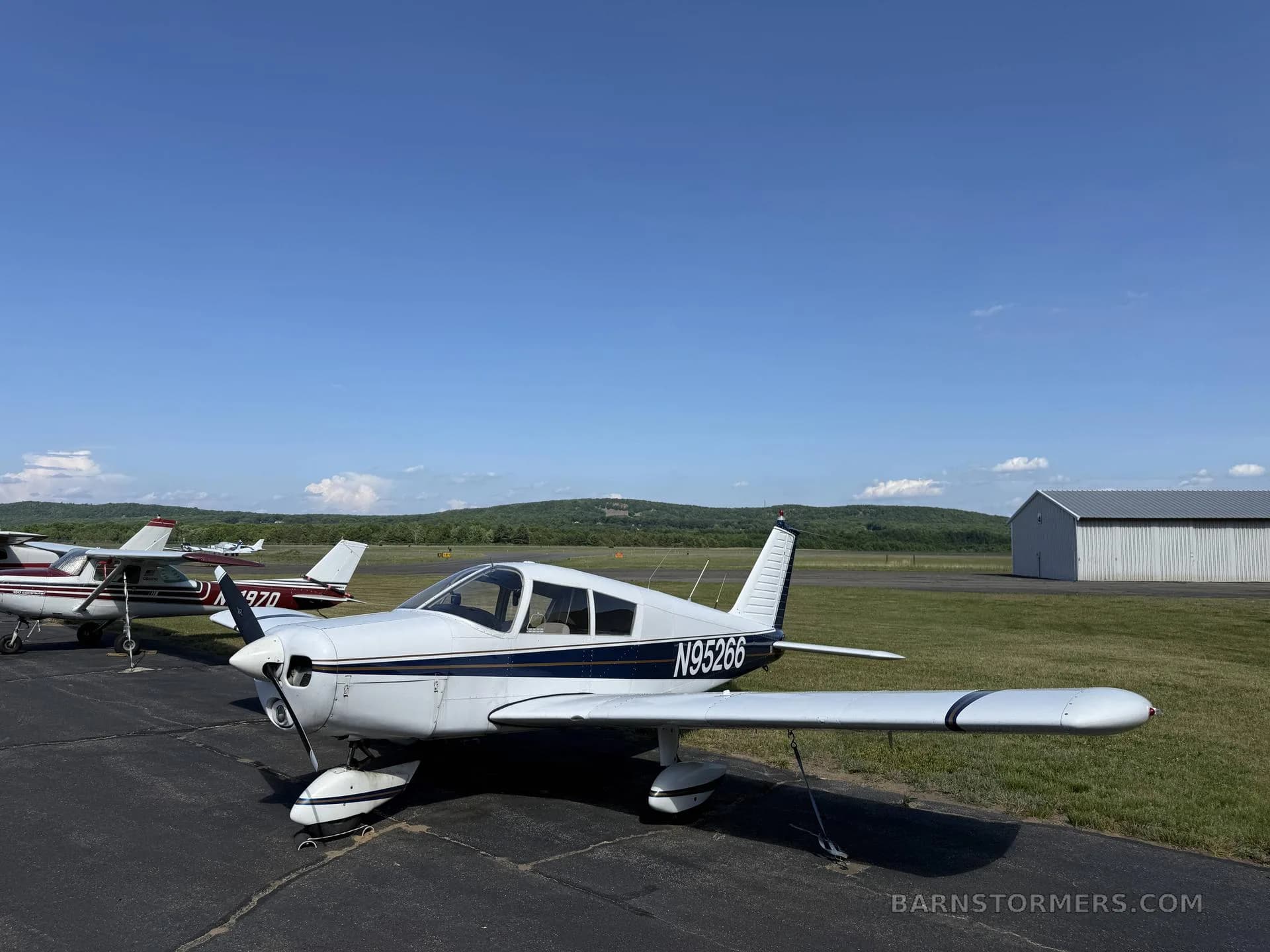 A white single-engine propeller aircraft (N95266) with blue stripes parked on an airport apron with other planes and a hangar.