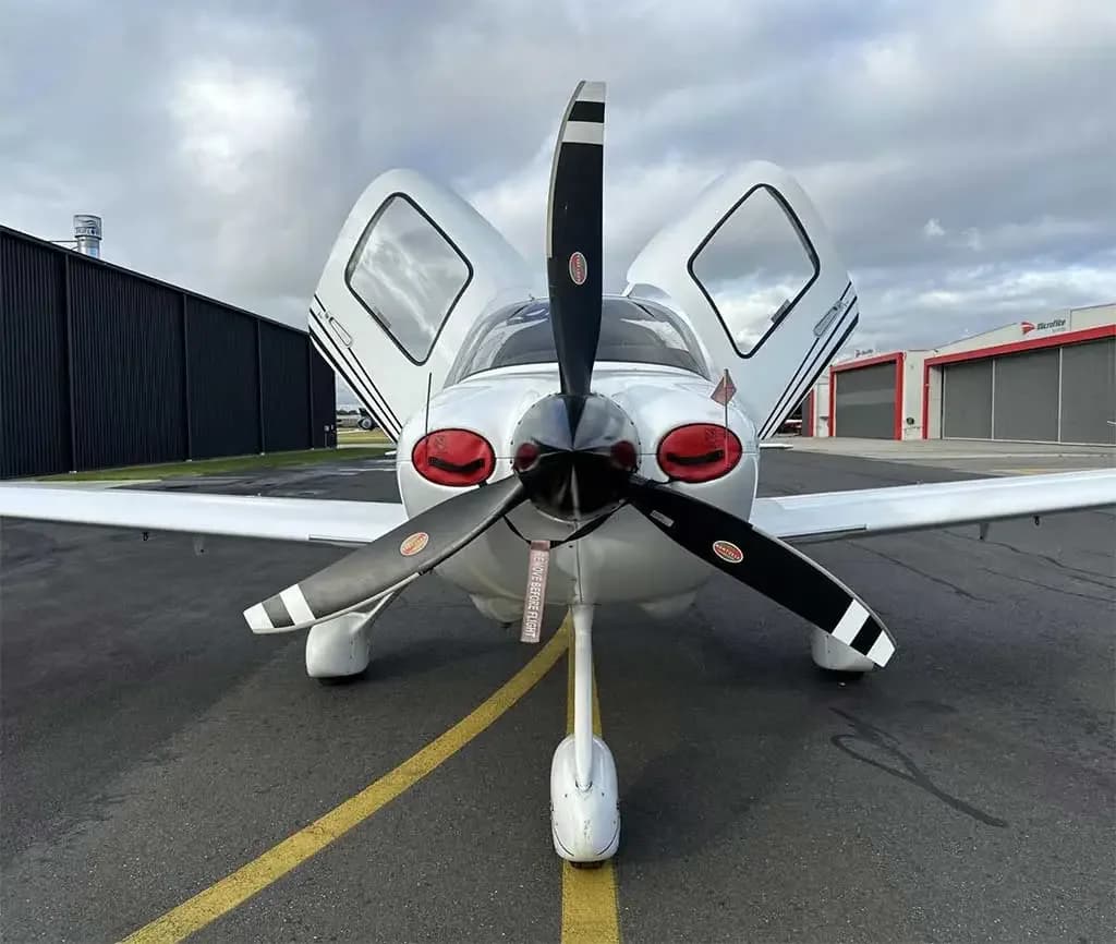 A head-on view of a white Cirrus SR22 aircraft with its gull-wing doors open, parked on a tarmac.