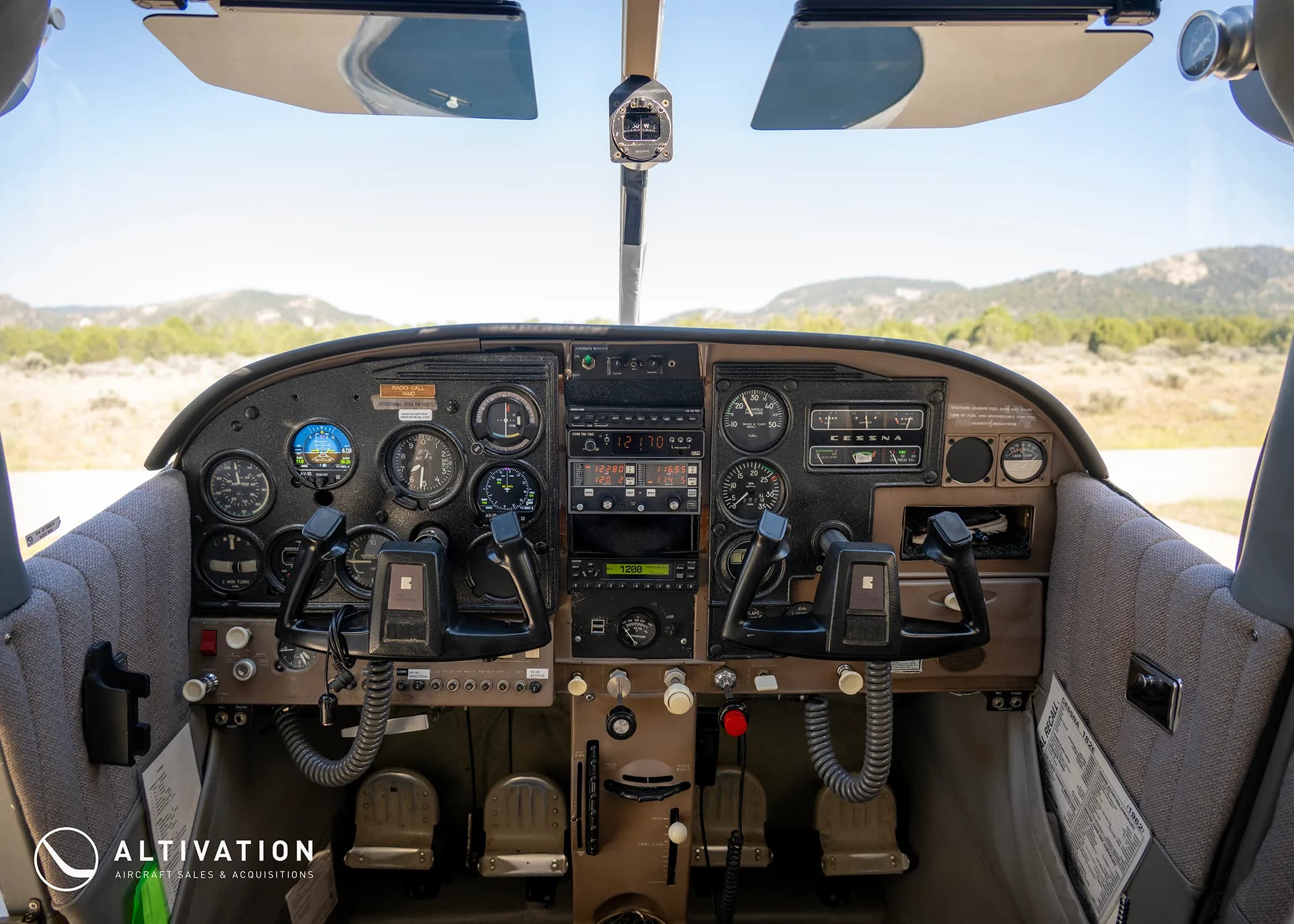 The cockpit of a Cessna aircraft, showing the instrument panel, yokes, and a view of mountains through the windshield.