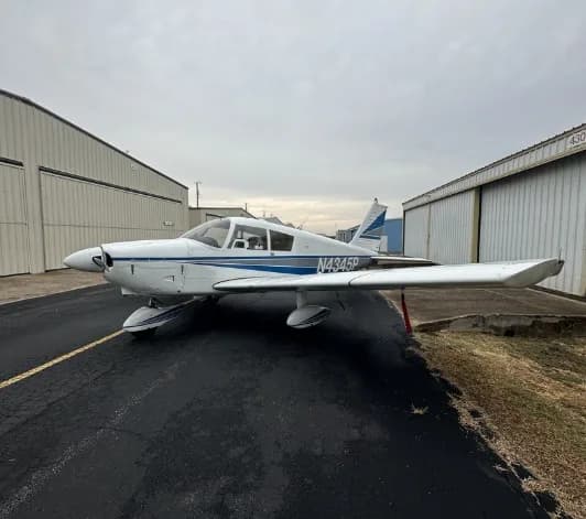 A white single-engine propeller aircraft with blue stripes (N4345P) is parked on asphalt between two hangars under an overcast sky.