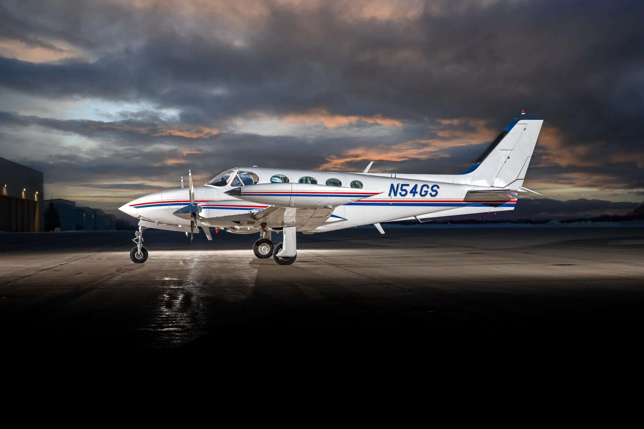 A white twin-engine propeller aircraft with blue and red stripes is parked on a tarmac at dusk under a dramatic cloudy sky.