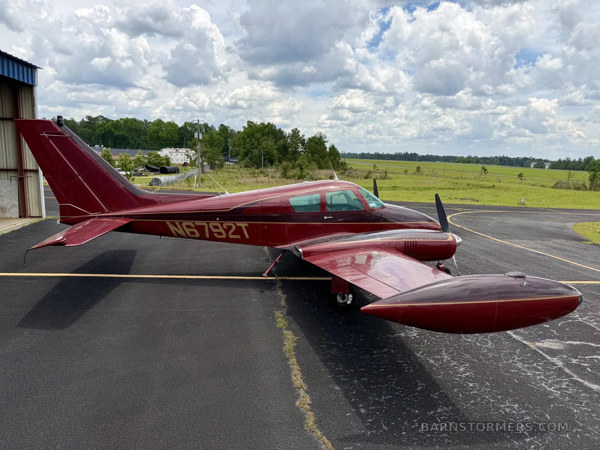 A red twin-engine aircraft, likely a Beechcraft Baron, with registration N6792T, parked on an asphalt apron with a grassy field in the background.