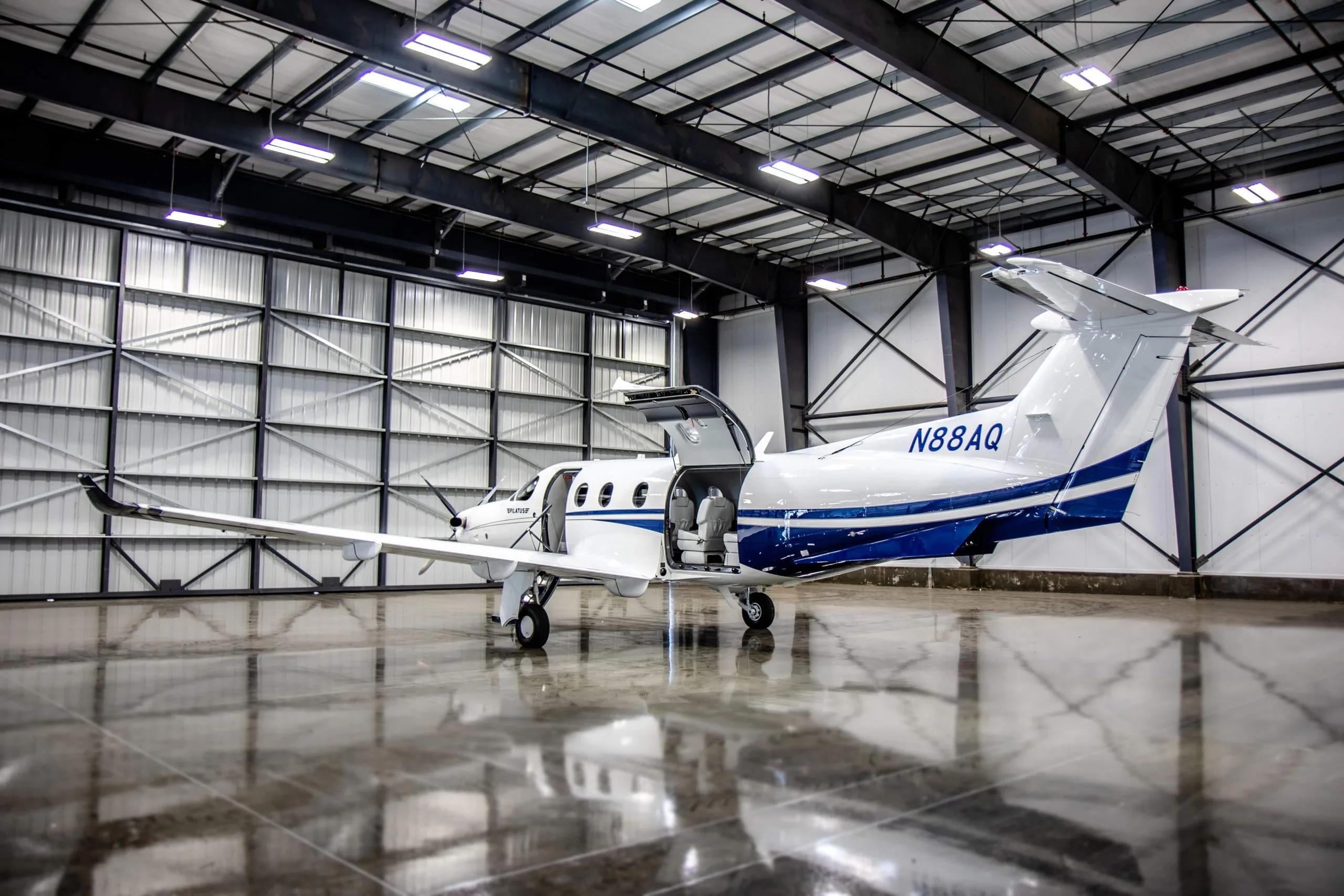 A white and blue Pilatus PC-12 aircraft with tail number N88AQ is parked in a hangar with its door open.