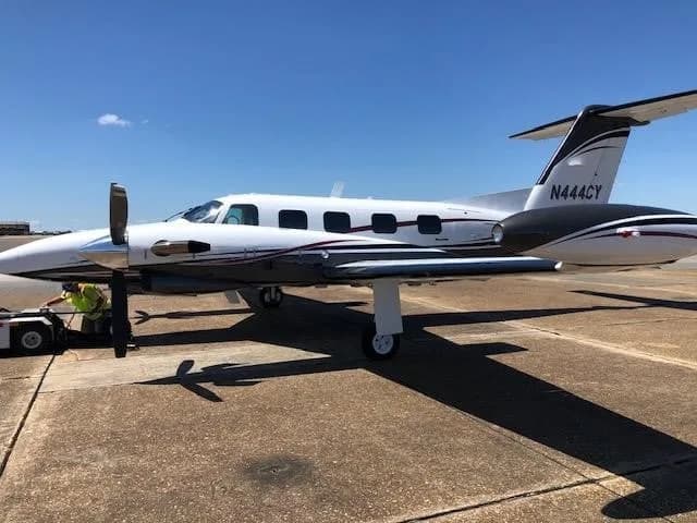A white and dark grey twin-engine turboprop aircraft with red pinstripes, tail number N444CY, is being towed on an airport tarmac under a clear blue sky.