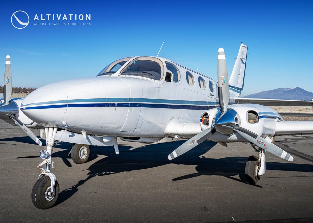 A white and blue twin-engine aircraft, registration N7719Q, sits on the tarmac in front of a hangar.
