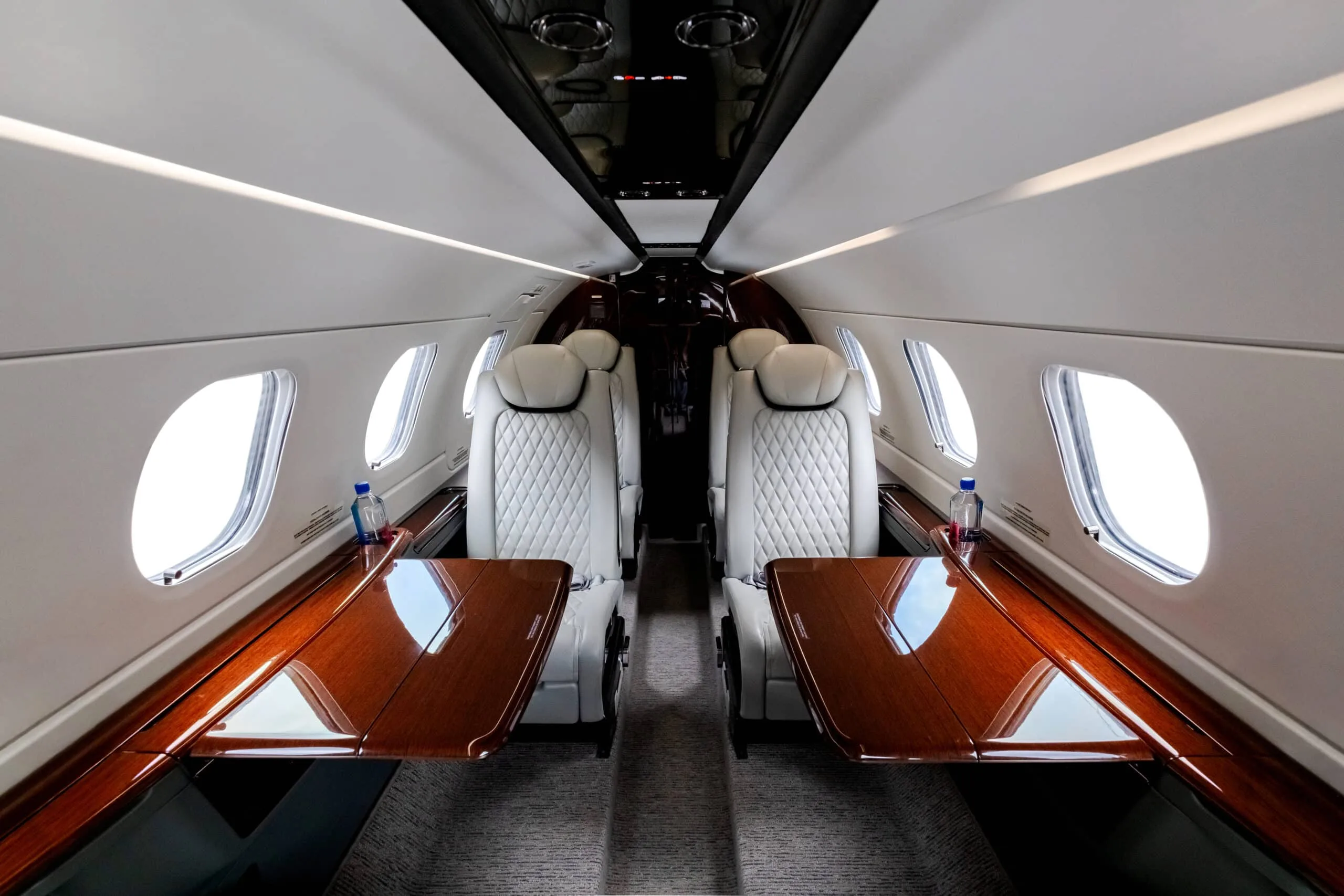 View down the aisle of the aircraft cabin with white leather seats and wood tables.