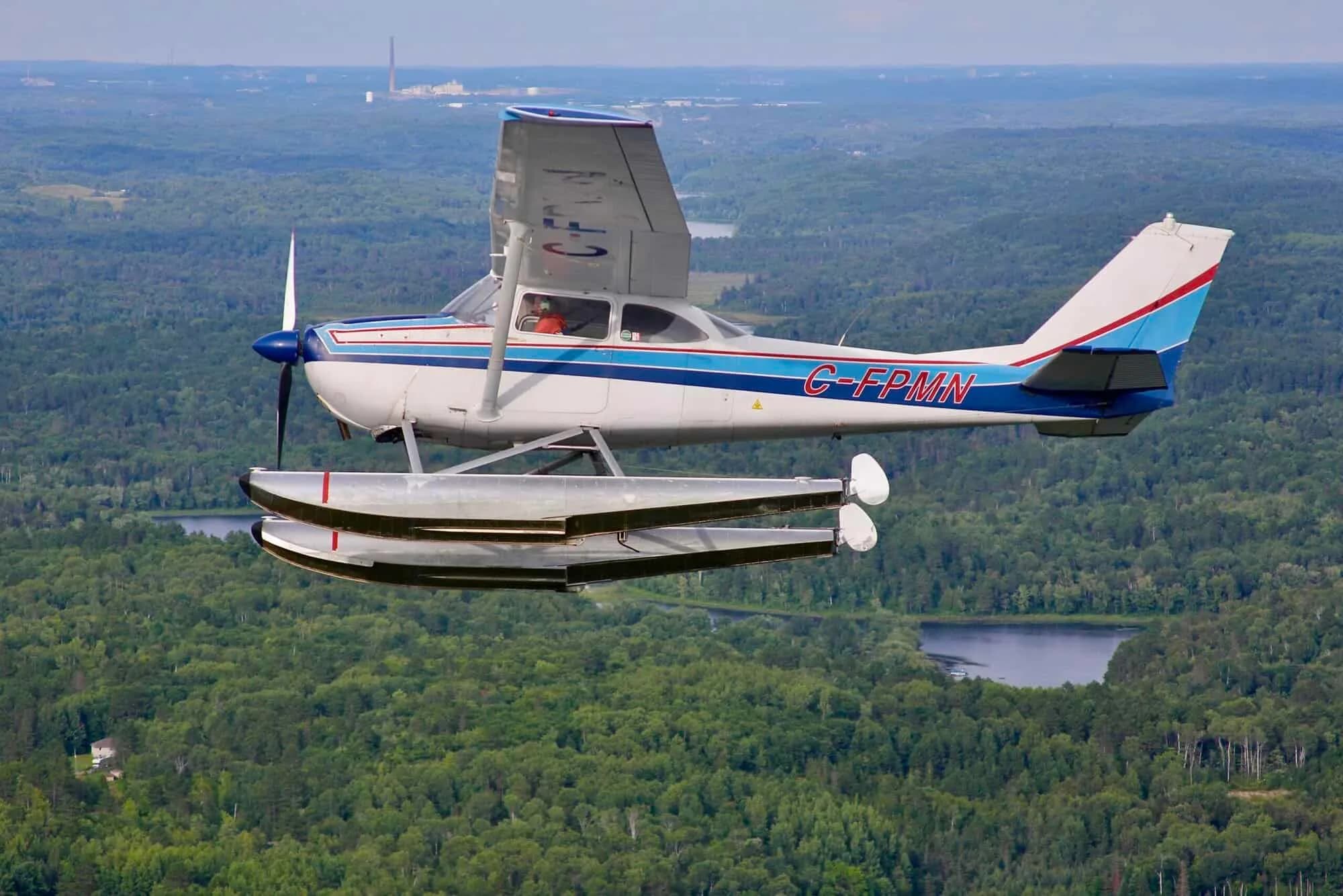 A white and blue Cessna 172 floatplane, registration C-FPMN, flies over a green forest and lakes.