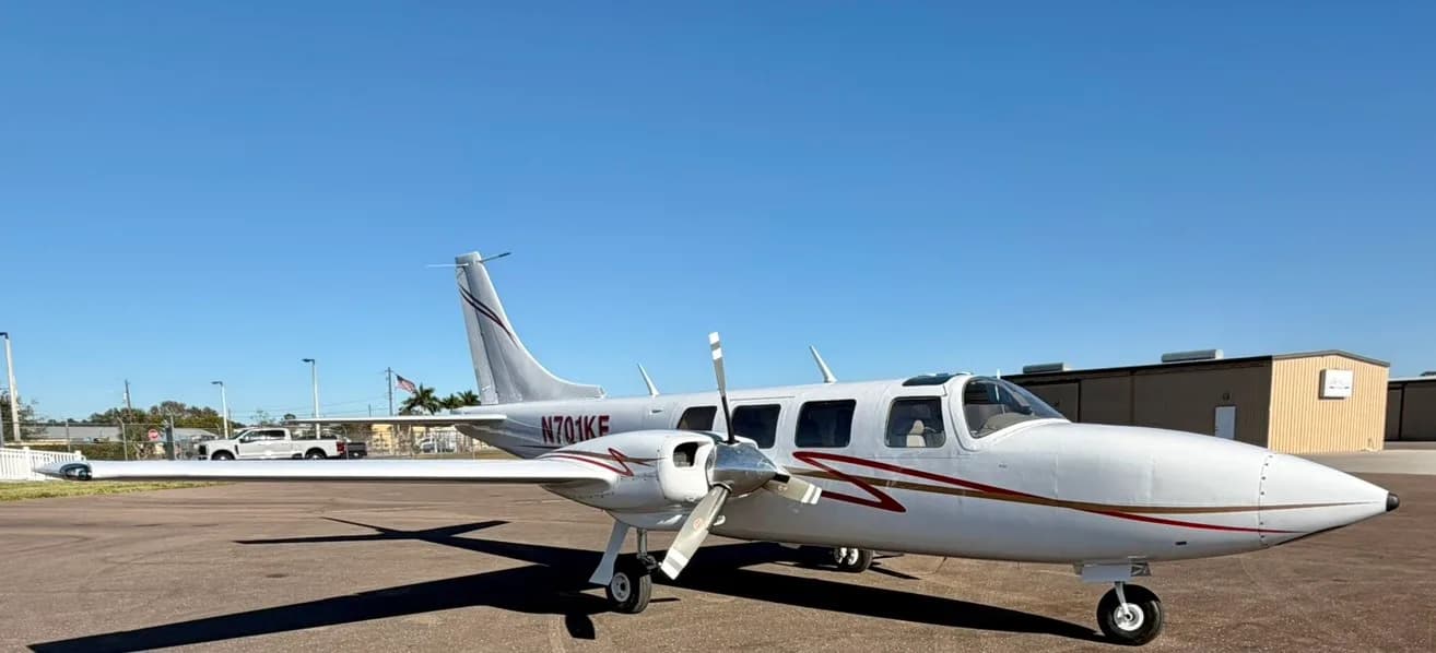 A white twin-engine propeller aircraft with red and gold stripes, registration N701KE, parked on an asphalt ramp with buildings in the background.