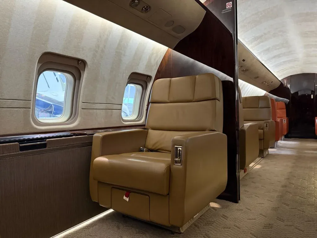 Multiple tan leather seats line the private jet cabin, with windows showing reflections of the outside.