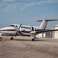 A white twin-turboprop aircraft with a dark red stripe is parked on an asphalt tarmac next to a hangar.