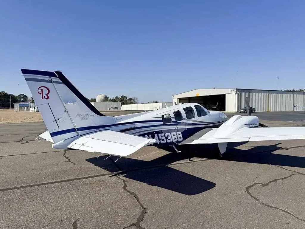 A white and blue Beechcraft Baron N453BB parked on an airfield with hangars in the background, viewed from the rear-left.