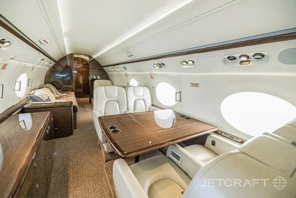 A view inside the aircraft cabin showing a dining area with a large wooden table and a lounge area further back.