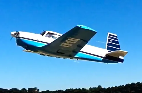 A white and blue single-engine propeller aircraft is flying against a clear blue sky, with trees visible below.