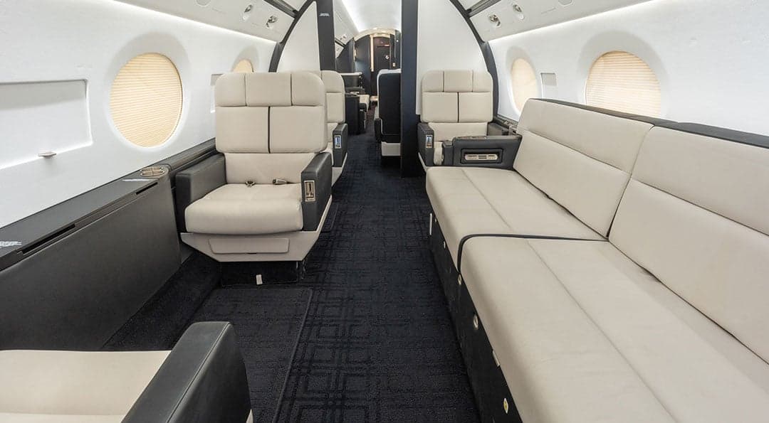 View of the Gulfstream G550 cabin with beige leather seats and dark carpeting.