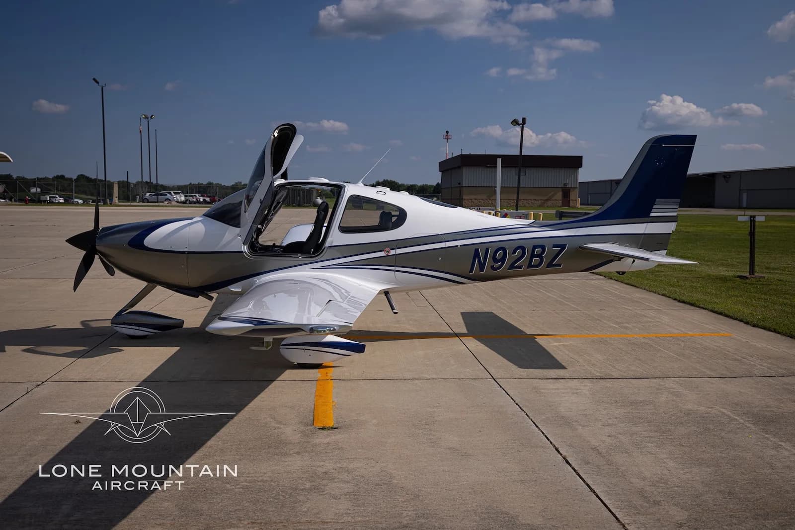 A side view of the white and silver Cirrus SR22 aircraft N92BZ with blue accents, parked on an airfield with its doors open.
