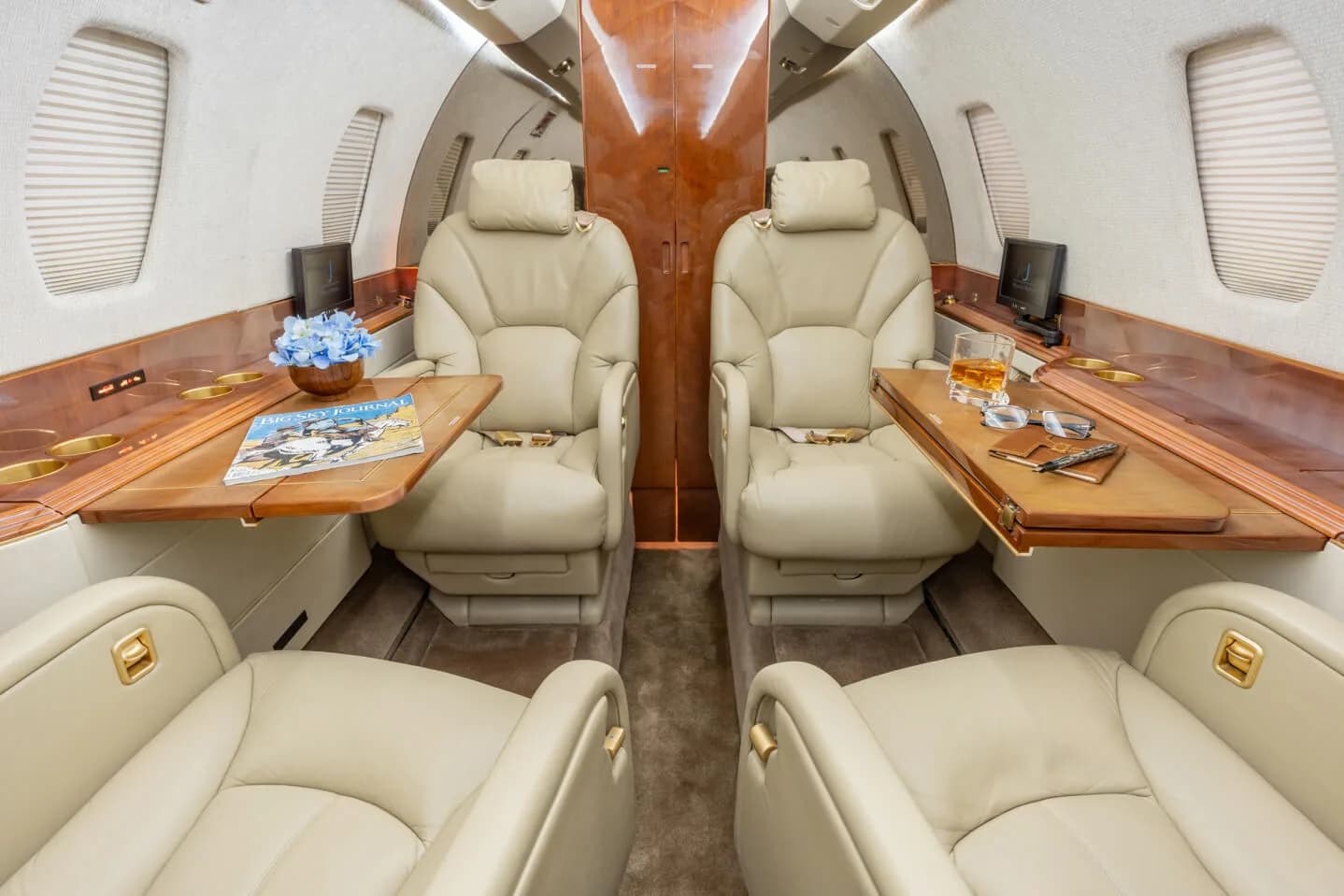 A view of the aircraft cabin with beige leather seats, wooden tables, a magazine, and a drink.