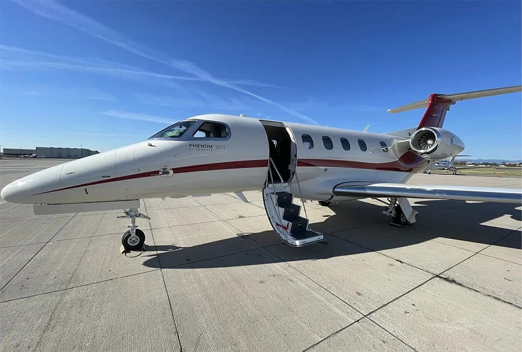 A white and red Embraer Phenom 300E private jet with its main cabin door open and stairs extended on the tarmac.