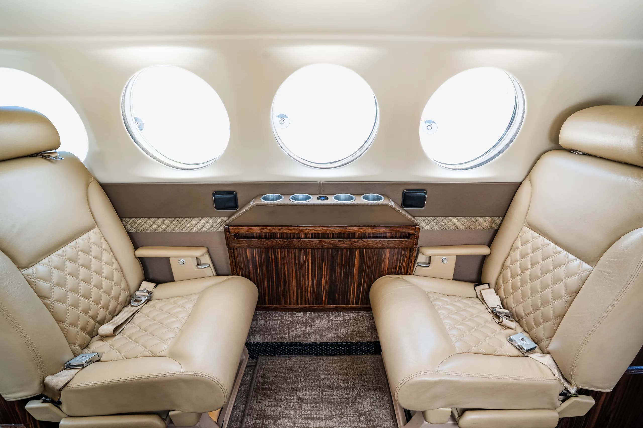 A close-up view of two beige leather aircraft cabin seats with a wooden console between them.