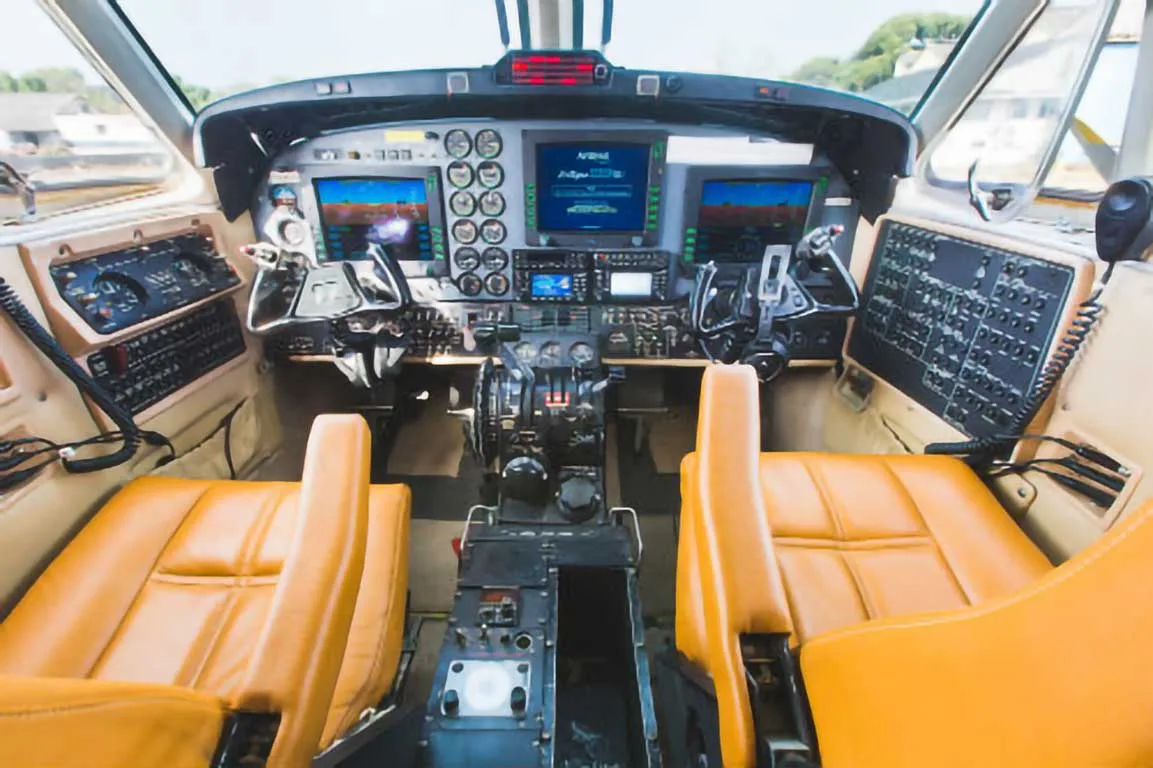 The cockpit of a twin-turboprop aircraft shows multiple digital displays, analog gauges, and tan leather seats.