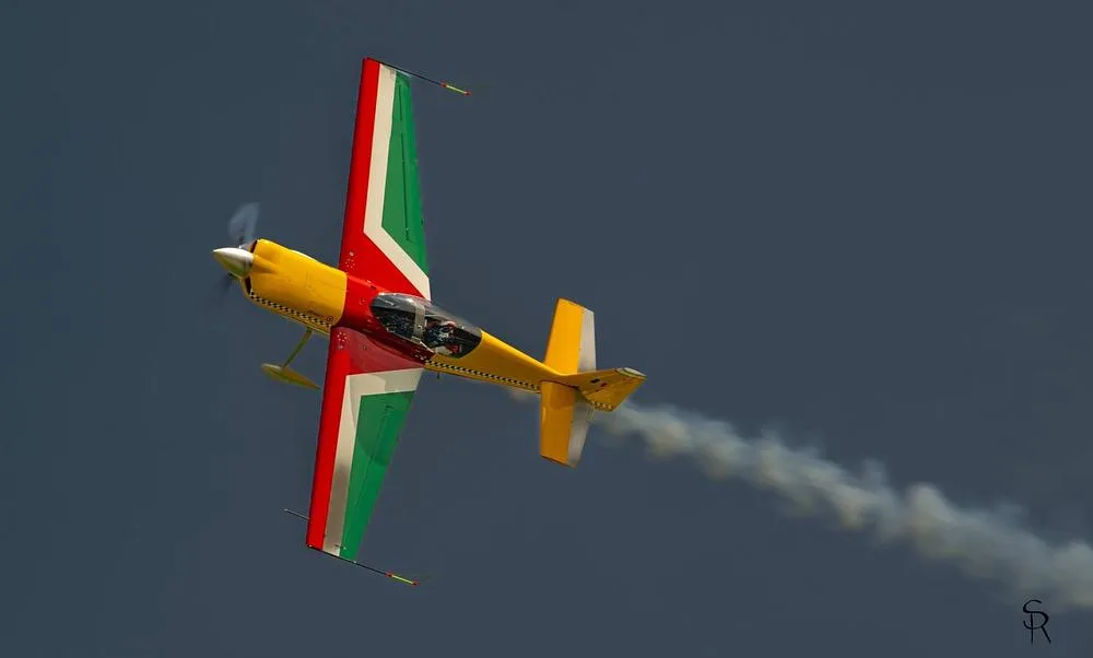 An aerobatic aircraft with red, green, and yellow livery performs a maneuver, leaving a smoke trail.