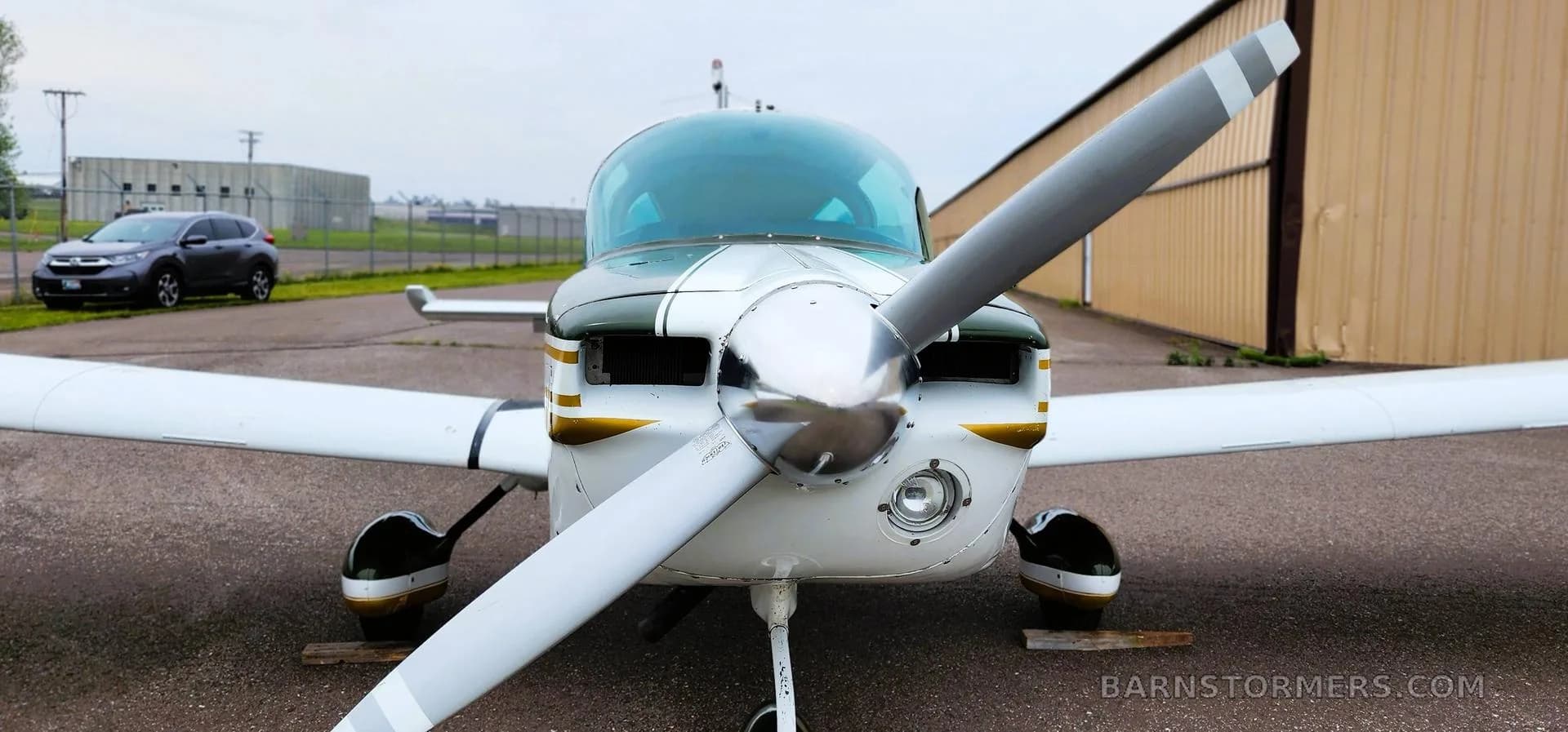 A head-on view of the propeller and nose of a white and dark green Grumman Tiger aircraft with gold stripes, parked on an asphalt ramp.