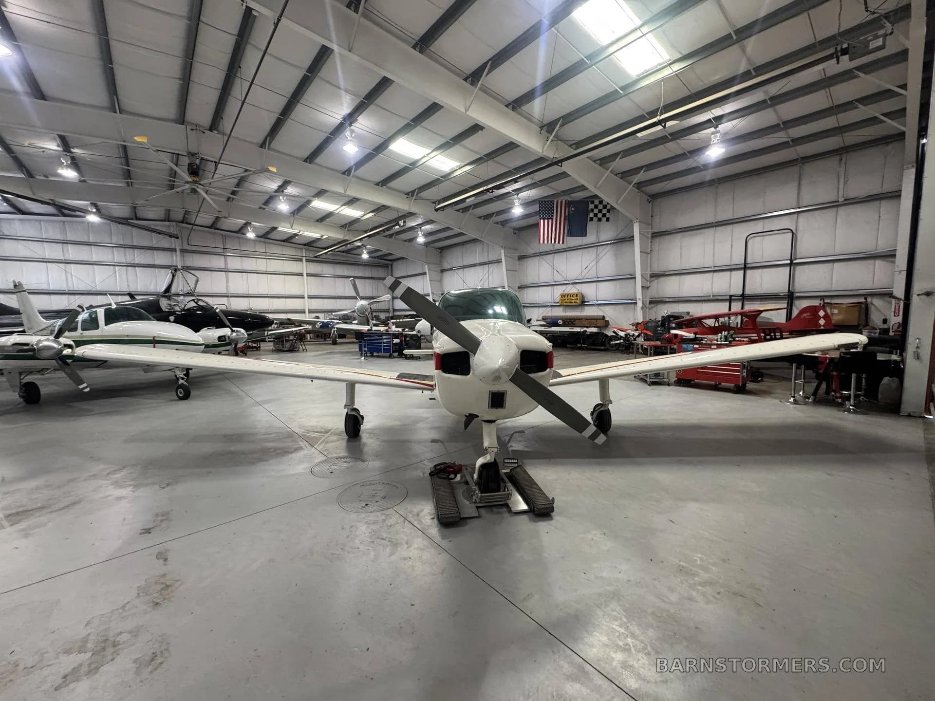 A white and red Beechcraft Sundowner N229FC is viewed head-on in a hangar, with other aircraft in the background.